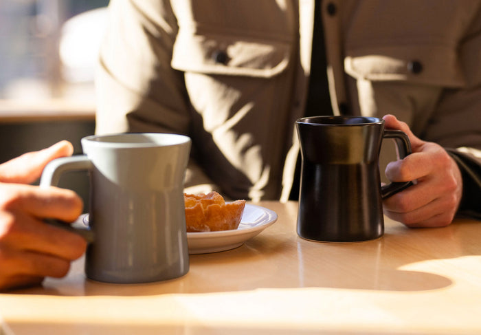 Two hands holding New Diner Mug handles at table 